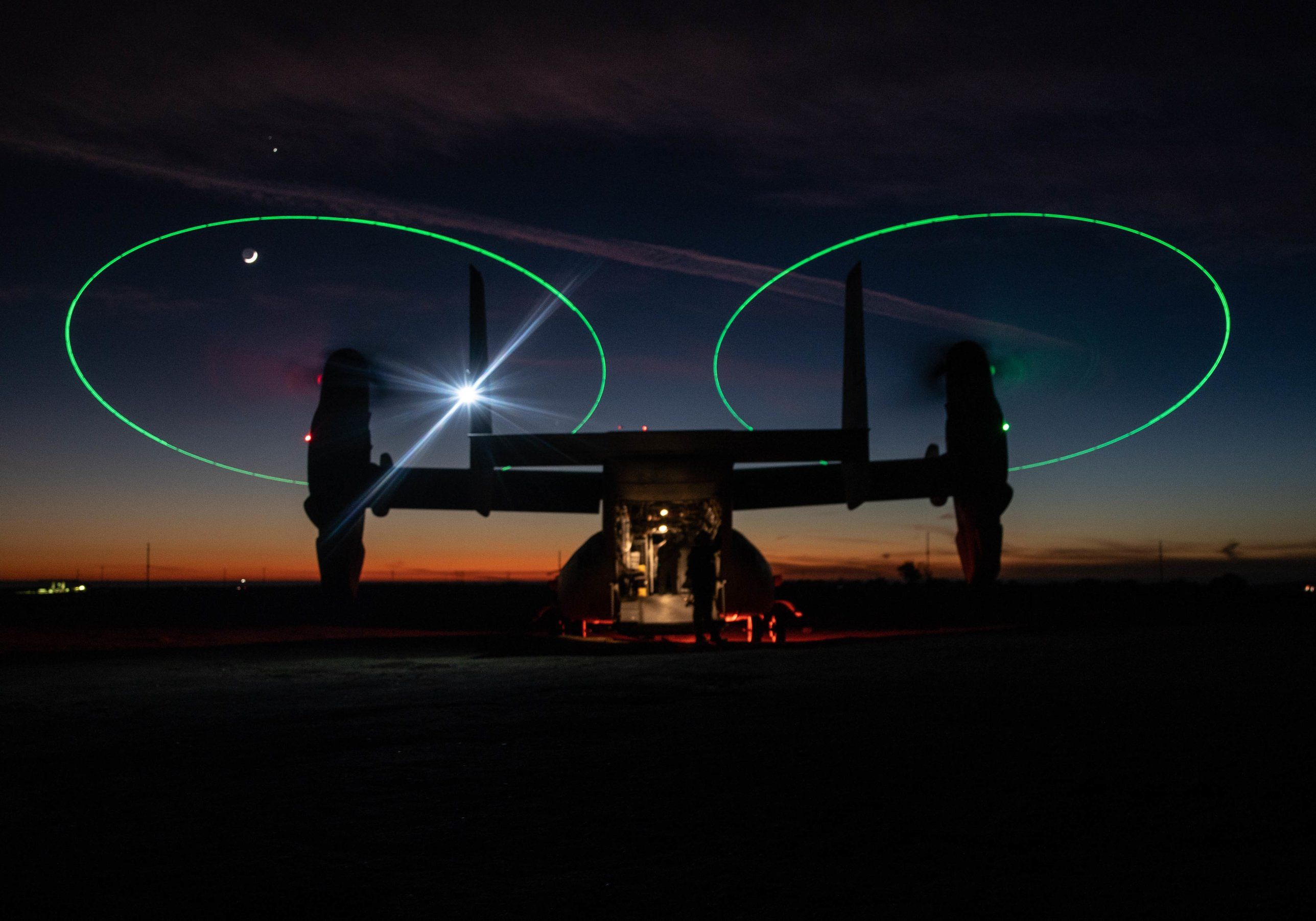 An MV-22 Osprey on the ground at last light, proprotors turning with green tip lights tracing two circles against the dusk sky